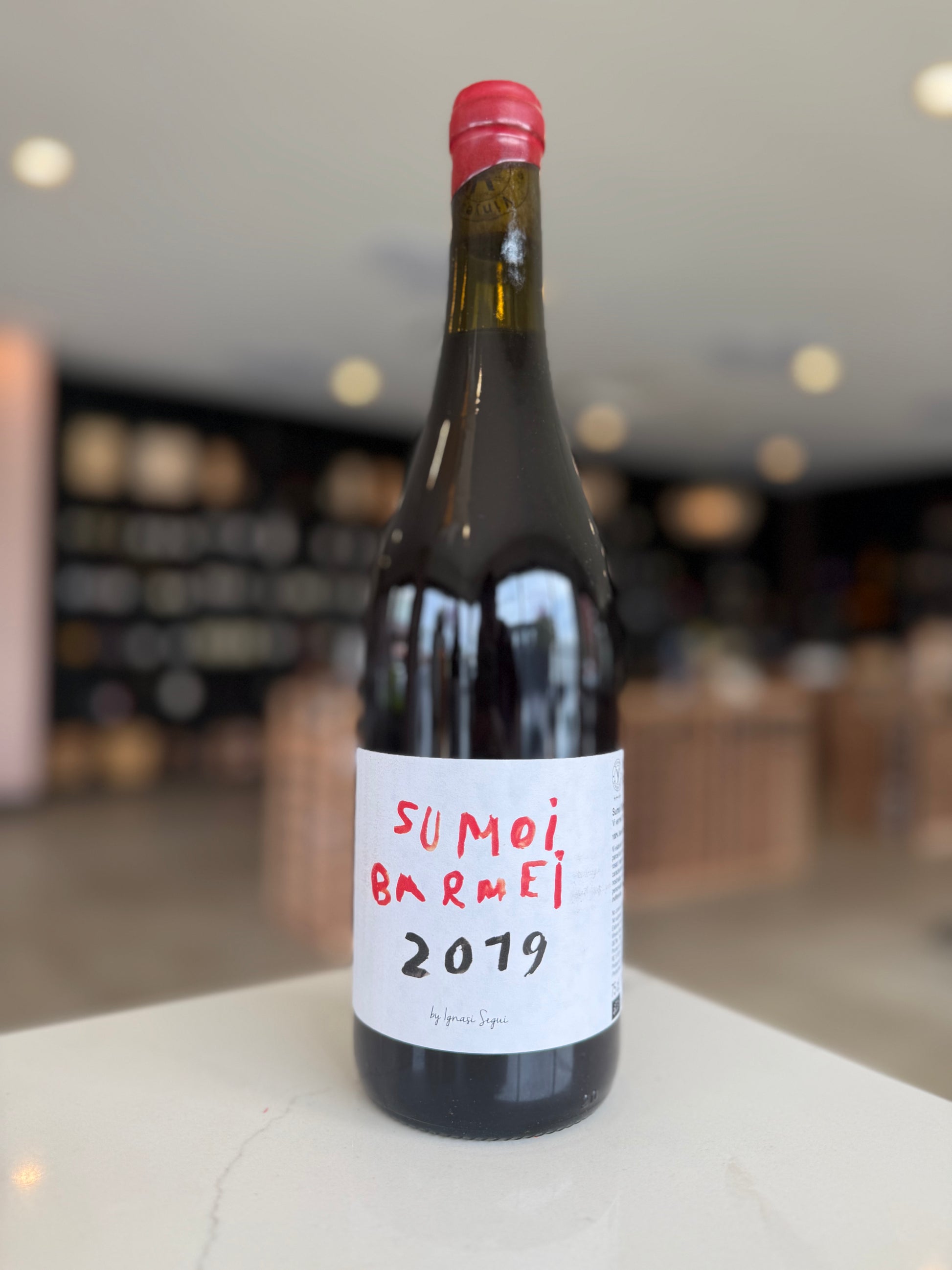 Wine bottle with a red cap and white label displaying 'Sumoi Barrei 2019' in a blurred indoor setting.